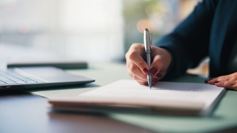 Osoba sedi za stolom i ručno vodi poslovne beleške pored laptopa, što prikazuje samostalno vođenje knjiga