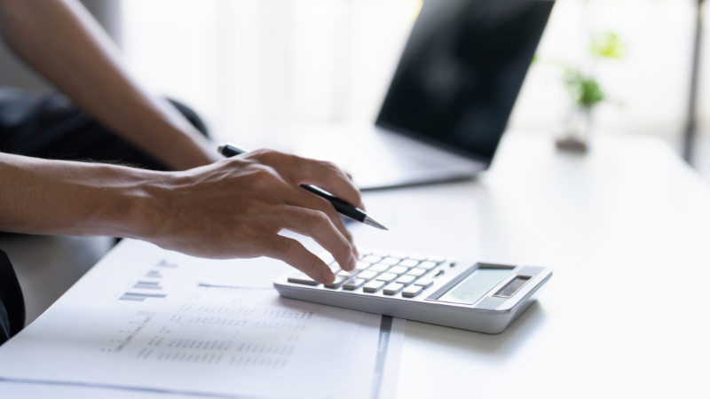 Osoba računa troškove na digitronu uz papire i laptop, što prikazuje samostalno vođenje finansija