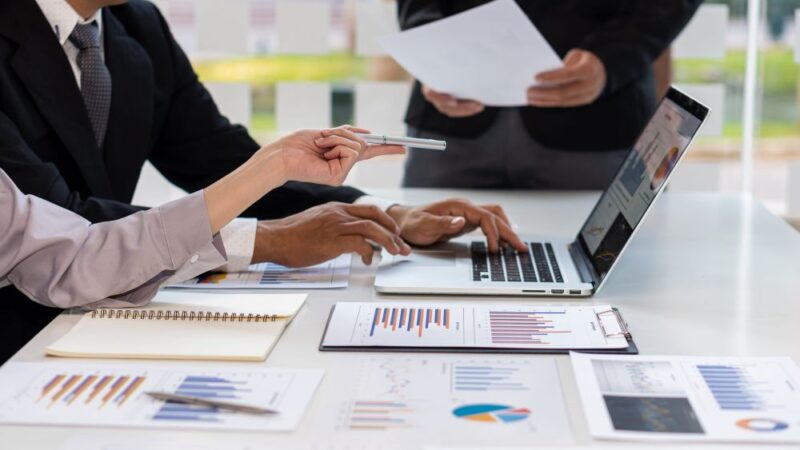 Zaposleni pregledaju papirnu i digitalnu poslovnu dokumentaciju za zajedničkim radnim stolom.